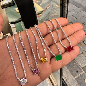 Puede incluir: Una colección de seis delicados collares, cada uno con un colgante rectangular de piedras preciosas en varios colores: transparente, rosa, amarillo, verde y rojo. Los colgantes están engarzados en una cadena plateada.