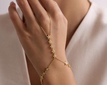 May include: A gold chain bracelet with a star design. The bracelet is connected to a finger ring, creating a hand chain.