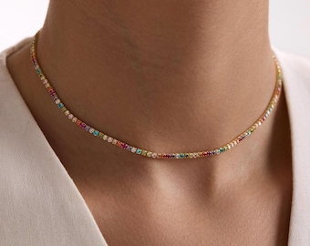 Könnte beinhalten: Eine goldfarbene Tennis-Halskette mit einer durchgehenden Reihe kleiner, mehrfarbiger Edelsteine in einem Regenbogenspektrum. Die Kette liegt eng am Hals an und präsentiert eine lebendige Anordnung roter, oranger, gelber, grüner, blauer und violetter Steine.