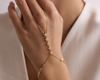 May include: A gold-toned hand chain featuring a delicate link bracelet connected to a finger ring by a chain adorned with multiple small star-shaped charms, each set with a tiny clear stone at its center, creating a celestial-inspired design.