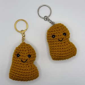 May include: Two crocheted brown poop emoji keychains with black eyes and a smiling mouth. Each keychain has a gold or silver metal ring.