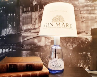 May include: A unique table lamp made from a Gin Mare gin bottle. The lamp features a white lampshade with the words "GIN MARE" and botanical illustrations. The bottle is clear with a blue base, and the background shows a cityscape.