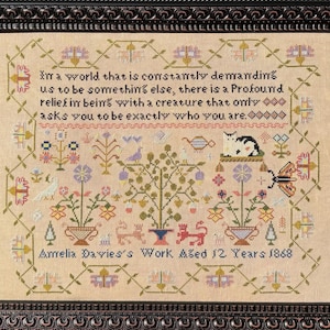 May include: A framed cross-stitch sampler on beige fabric featuring a floral border, a central tree, a cat, birds, and butterflies. The text reads: In a world that is constantly demanding us to be something else, there is a profound relief in being with a creature that only asks you to be exactly who you are. Amelia Davies's Work Aged 12 Years 1868.