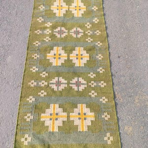 Pode incluir: Um tapete verde, amarelo e branco com franjas nas extremidades. O tapete tem um design geométrico com padrões repetidos.