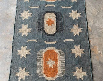 May include: A blue rectangular rug with a star and geometric pattern. The rug features repeating star designs in white and orange geometric shapes. The rug has a fringed edge and is suitable for a living room or bedroom.