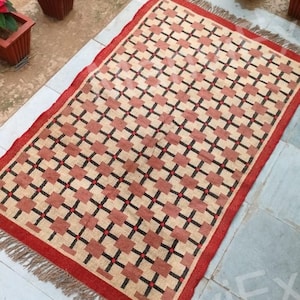 May include: A red, black, and cream patterned rug with fringe edges. The rug is laid out on a tiled floor.