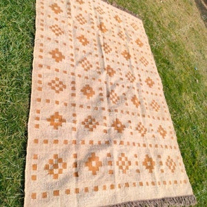 May include: A rectangular beige rug with a repeating geometric pattern in shades of brown. The rug has a fringed edge and is displayed on a green grassy surface. The pattern includes squares and diamond shapes.