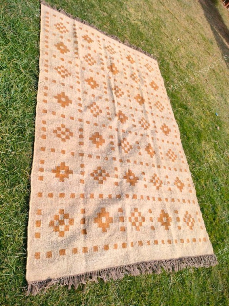 May include: A rectangular beige rug with a repeating geometric pattern in shades of brown. The rug has a fringed edge and is displayed on a green grassy surface. The pattern includes squares and diamond shapes.
