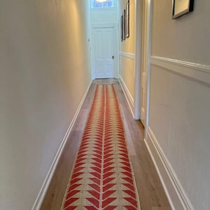 Può includere: Un corridoio lungo e stretto con pareti bianche e pavimento in legno. Un tappeto con motivo rosso e beige corre al centro del corridoio. Una porta bianca si trova alla fine del corridoio e un lampadario pende dal soffitto.