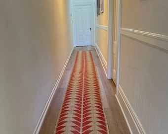 Puede incluir: Un pasillo largo y estrecho con paredes blancas y suelo de madera. Una alfombra con estampado rojo y beige recorre el centro del pasillo. Una puerta blanca se encuentra al final del pasillo, y una lámpara cuelga del techo.