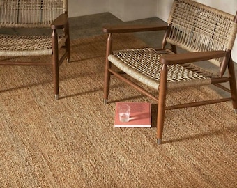 Puede incluir: Dos sillones de madera con asientos y respaldos tejidos sobre una alfombra de yute tejida. Un vaso de agua y un libro descansan sobre la alfombra entre las sillas. Las sillas tienen un diseño moderno de mediados de siglo.