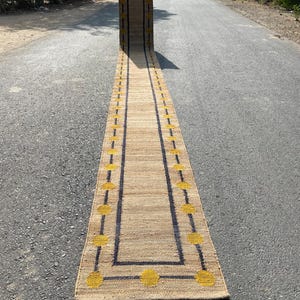 Puede incluir: Una alfombra larga y estrecha de fibra natural con un borde negro y círculos amarillos. La alfombra está colocada en un camino pavimentado.