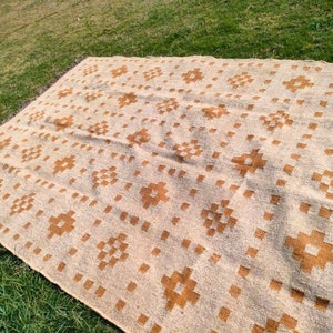 May include: A large, rectangular beige rug with a repeating pattern of brown floral and square designs. The rug is laid out on green grass, showcasing its size and texture. The edges have a fringed detail.