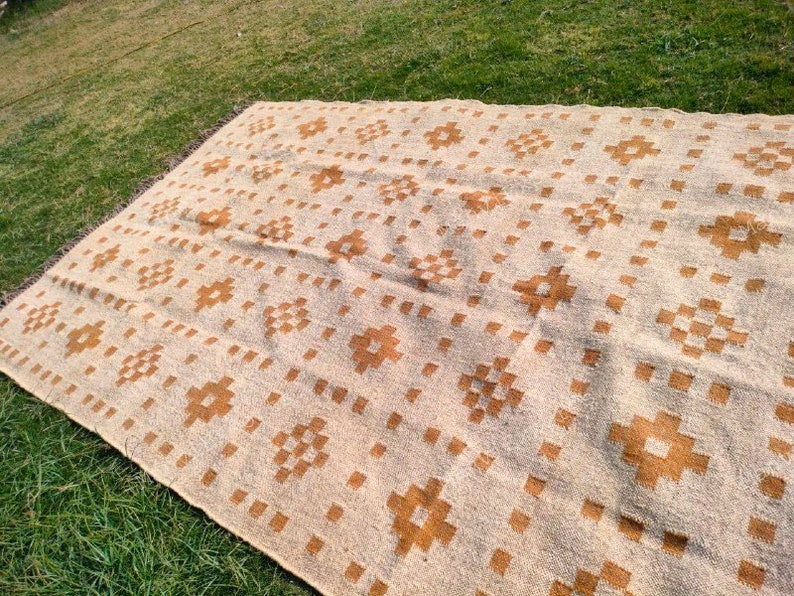 May include: A large, rectangular beige rug with a repeating pattern of brown floral and square designs. The rug is laid out on green grass, showcasing its size and texture. The edges have a fringed detail.