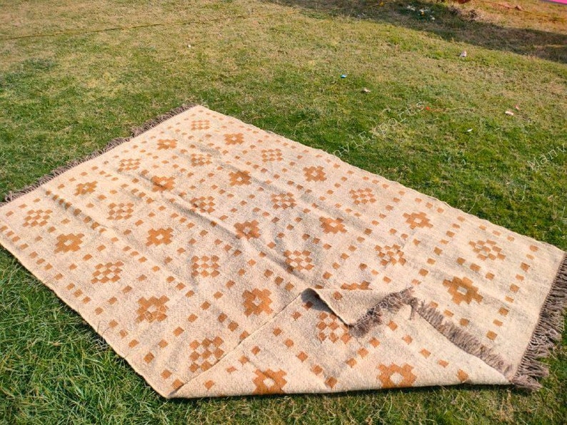 May include: A beige and brown patterned rug with a geometric design. The rug has a rectangular shape and fringed edges. The pattern features repeating square and flower-like motifs. The rug is laid out on a green grassy surface.