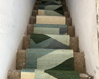 May include: A set of stair treads with a geometric pattern in shades of green, blue, and beige. The treads are made of a natural fiber material, such as jute or sisal. The treads are laid out on a set of stairs in a home.