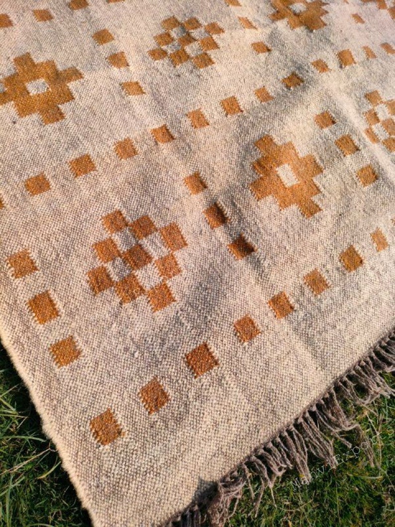 May include: Beige and brown woven rug with a geometric pattern. The rug features a repeating design of brown squares and cross-like shapes. The edges have a fringe detail.