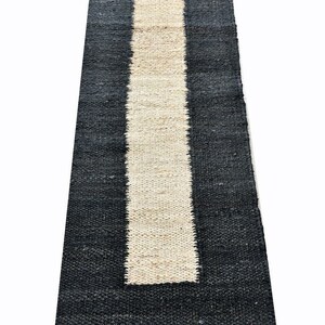 May include: A rectangular rug with a black border and a central, vertical stripe in a natural, light beige color. The rug has a woven texture, and the design is simple and modern. The rug is suitable for indoor use.