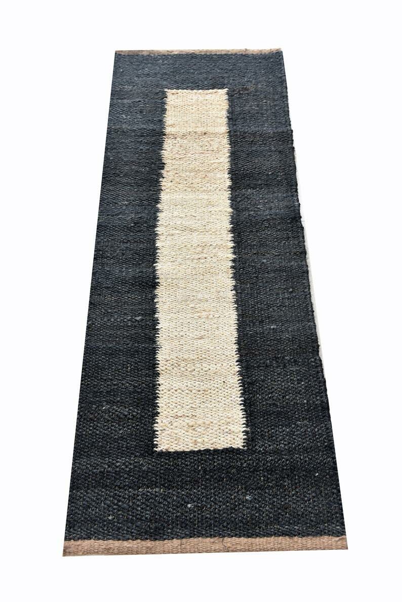 May include: A rectangular rug with a black border and a central, vertical stripe in a natural, light beige color. The rug has a woven texture, and the design is simple and modern. The rug is suitable for indoor use.