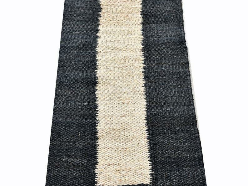 May include: A rectangular rug with a central cream-colored stripe flanked by black stripes. The rug has a woven texture and is likely made of natural fibers. This rug is suitable for home decor.