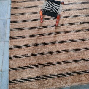May include: A natural fiber rug with black and brown stripes. A small wooden stool with a black and white patterned seat sits on the rug.