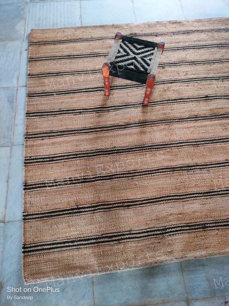 May include: A natural fiber rug with black and brown stripes. A small wooden stool with a black and white patterned seat sits on the rug.