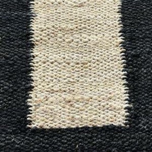 May include: A rectangular rug with a woven texture. The rug features a central vertical stripe in a natural beige color, framed by a solid black border. The rug's design is simple and modern, suitable for various interior styles.