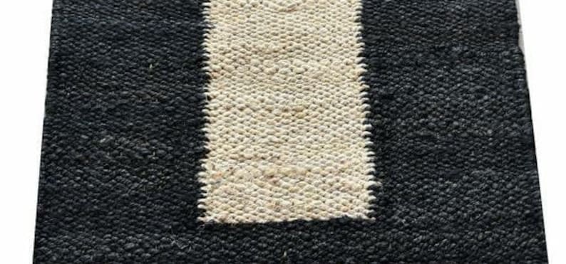 May include: A rectangular rug with a woven texture. The rug features a central vertical stripe in a natural beige color, framed by a solid black border. The rug's design is simple and modern, suitable for various interior styles.