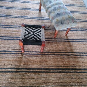 May include: Two wooden stools with fabric seats. The larger stool has a light blue fabric with a white and red geometric pattern. The smaller stool has a black and white geometric pattern. Both stools are sitting on a striped rug with brown and black stripes.