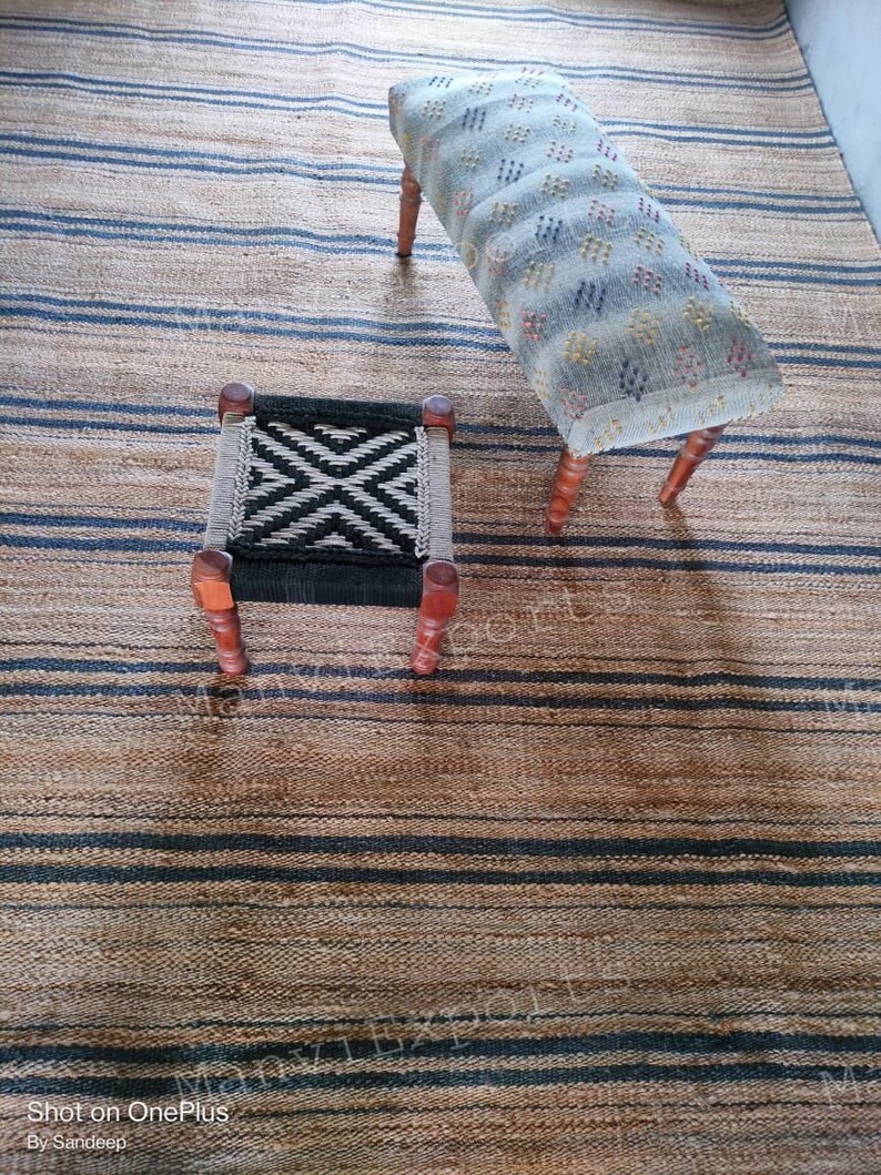 May include: Two wooden stools with fabric seats. The larger stool has a light blue fabric with a white and red geometric pattern. The smaller stool has a black and white geometric pattern. Both stools are sitting on a striped rug with brown and black stripes.