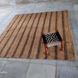 May include: A natural fiber rug with black and brown stripes. The rug is made of jute and is perfect for adding a touch of natural beauty to any room. A small wooden stool with a black and white woven seat sits on the rug.