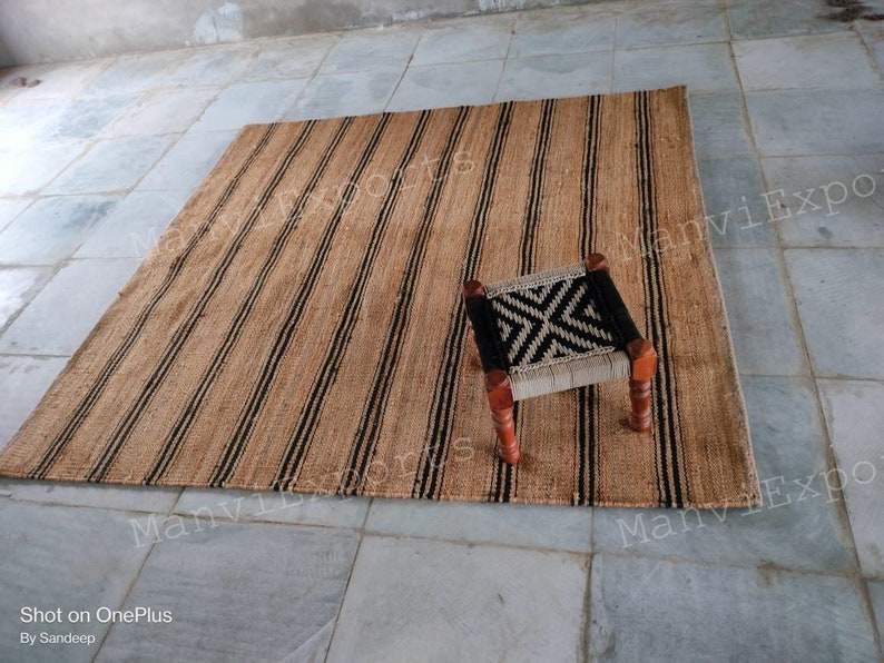May include: A natural fiber rug with black and brown stripes. The rug is made of jute and is perfect for adding a touch of natural beauty to any room. A small wooden stool with a black and white woven seat sits on the rug.