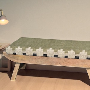 Pode incluir: Um banco de madeira com uma almofada estampada em verde e branco. Uma pequena mesa lateral e uma lâmpada dourada com uma abajur rosa claro estão ao lado do banco. A lâmpada está ligada.