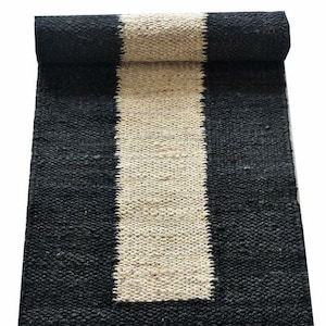 May include: A rolled-up jute runner rug with a black border and a central stripe of natural beige. The rug's woven texture is visible, and it is set against a white background. This rug is suitable for home decor.