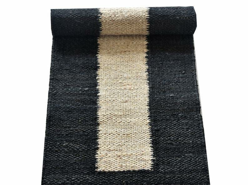May include: A rolled-up jute runner rug with a black border and a central stripe of natural beige. The rug's woven texture is visible, and it is set against a white background. This rug is suitable for home decor.