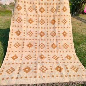 Puede incluir: Alfombra beige y marrón con diseño geométrico. La alfombra presenta patrones repetidos de diamantes y cuadrados marrones sobre un fondo beige. La alfombra tiene un borde con flecos y se exhibe al aire libre sobre hierba.
