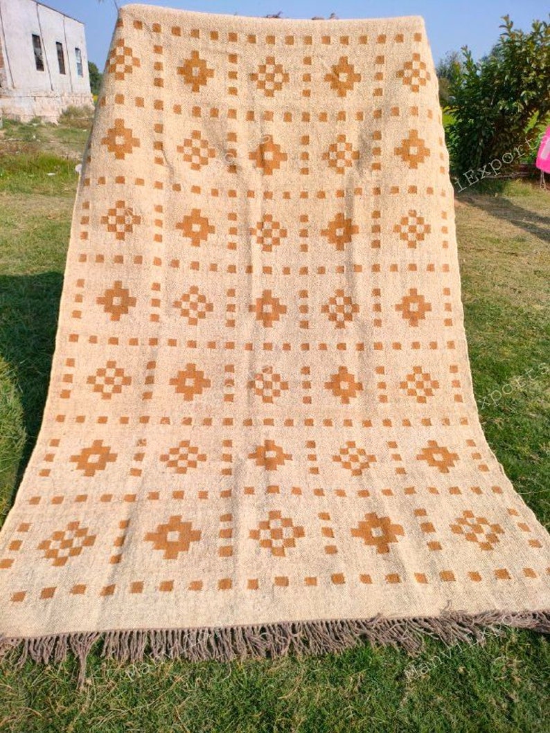 May include: Beige and brown patterned rug with a geometric design. The rug features repeating brown diamond and square patterns on a beige background. The rug has a fringed edge and is displayed outdoors on grass.