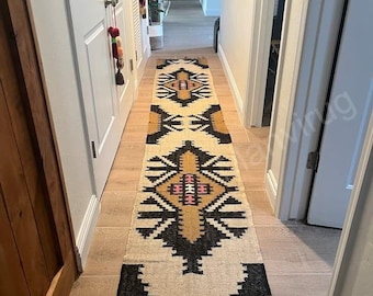 Puede incluir: Una alfombra larga y estrecha con un patrón geométrico en negro, blanco, marrón y rosa. La alfombra está colocada en un pasillo con paredes blancas y suelo de madera.