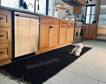May include: A black jute rug with frayed edges in a kitchen with wood cabinets and a white dog sitting on the rug.