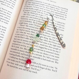 May include: A handmade beaded bookmark with a silver floral design and a mushroom charm. The bookmark features colorful beads in shades of green, yellow, and red, perfect for marking pages in a book.