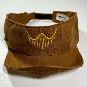 May include: A brown visor with a curved brim and a black adjustable strap. The visor features a gold embroidered design on the front. The visor is made of a durable fabric and is designed to shield the eyes from the sun.