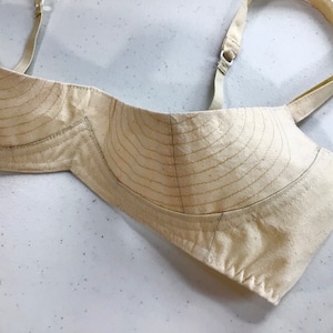 May include: A beige bra cup with a pattern of stitched lines. The bra cup is made of a soft, lightweight fabric.