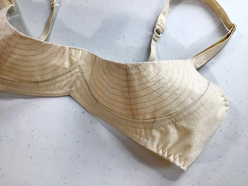 May include: A beige bra cup with a pattern of stitched lines. The bra cup is made of a soft, lightweight fabric.