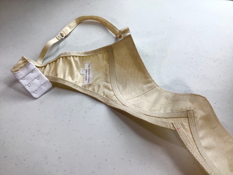 May include: A beige bra with a white label that reads "The Wilder Collection, UK Handmade". The bra has a hook and eye closure and is made of a soft, natural fabric.