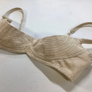 May include: A beige cotton bra with adjustable straps. The cups are lined with horizontal stitching.