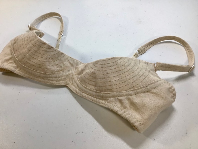 May include: A beige cotton bra with adjustable straps. The cups are lined with horizontal stitching.
