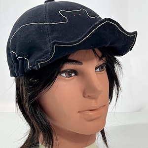 May include: Black cap with a cat outline design. The cat is outlined in white stitching and has red eyes. The cap has a curved brim and is made of a dark fabric.