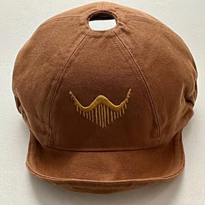 May include: A brown cap with a small brim and a circular opening at the top. The cap features a gold embroidered design on the front. The cap is made of a soft, textured fabric.