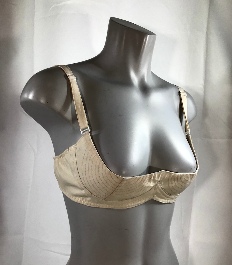 May include: A beige bra with a unique design featuring a curved, padded cup. The bra has adjustable straps and a smooth, satin-like finish.