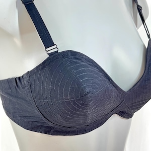 May include: A black bra with adjustable straps and a structured design. The bra features a series of concentric stitched lines on the cups, creating a textured effect. The straps and band are also black, with metal hardware.